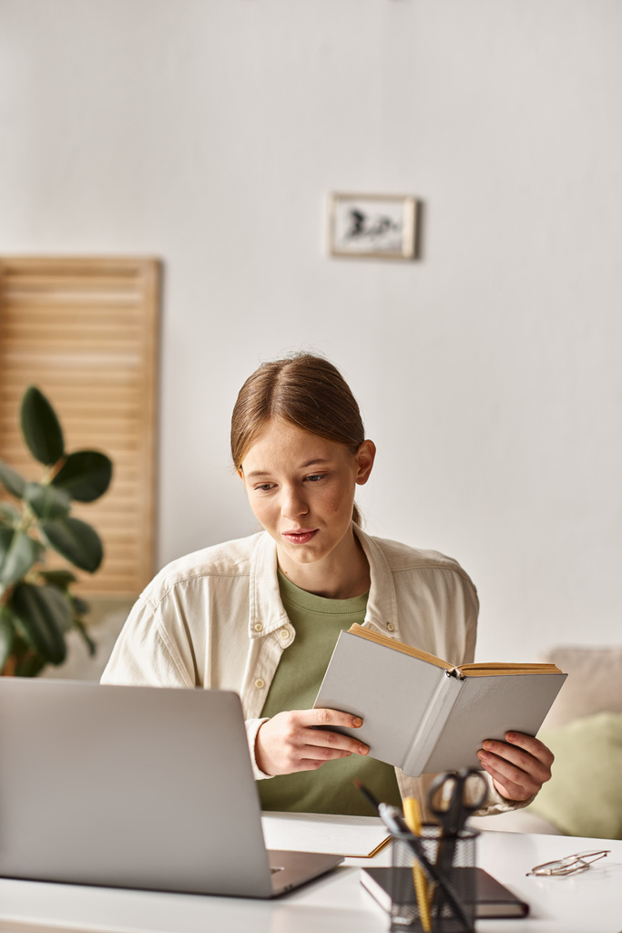zaneprázdněná teenagerka drží učebnici a sedí doma před laptopem, e-learning - Fotografie, Obrázek
