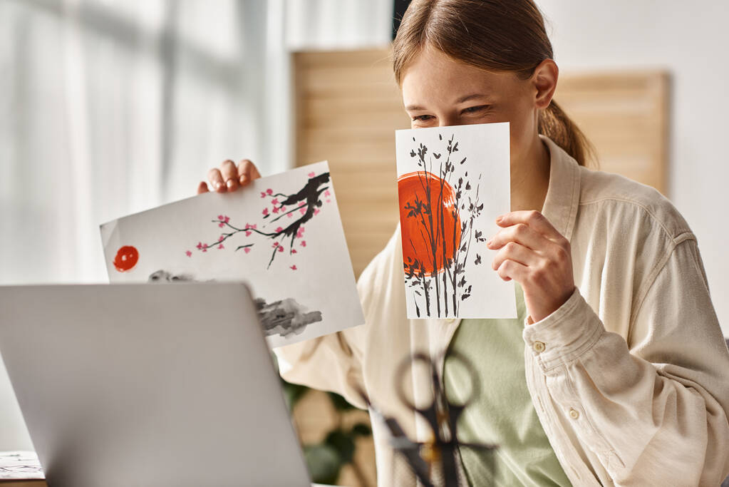 Ragazza adolescente che mostra le sue opere d'arte mentre copre il viso e guardando il suo computer portatile, classe d'arte online - Foto, immagini