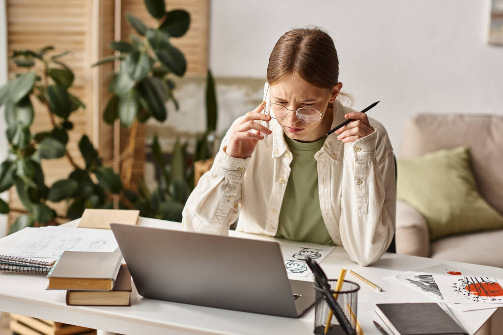 девочка-подросток разговаривает на смартфоне и смотрит на ноутбук рядом со своим рисунком на столе - Фото, изображение