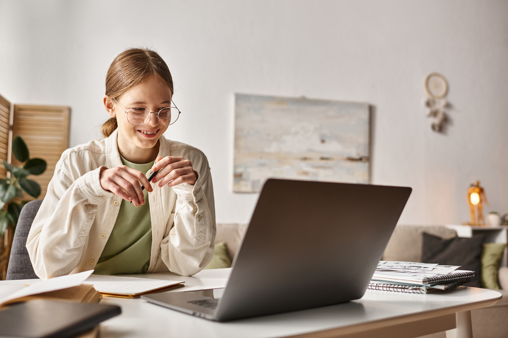 χαρούμενη έφηβη με γυαλιά κρατώντας στυλό και γράφοντας κατά τη διάρκεια του online μαθήματος στο laptop, κρατώντας σημειώσεις - Φωτογραφία, εικόνα