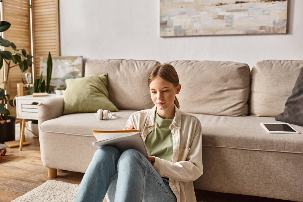 Ненавязчивая девочка-подросток читает свой ноутбук возле дивана с наушниками и цифровым планшетом рядом - Фото, изображение