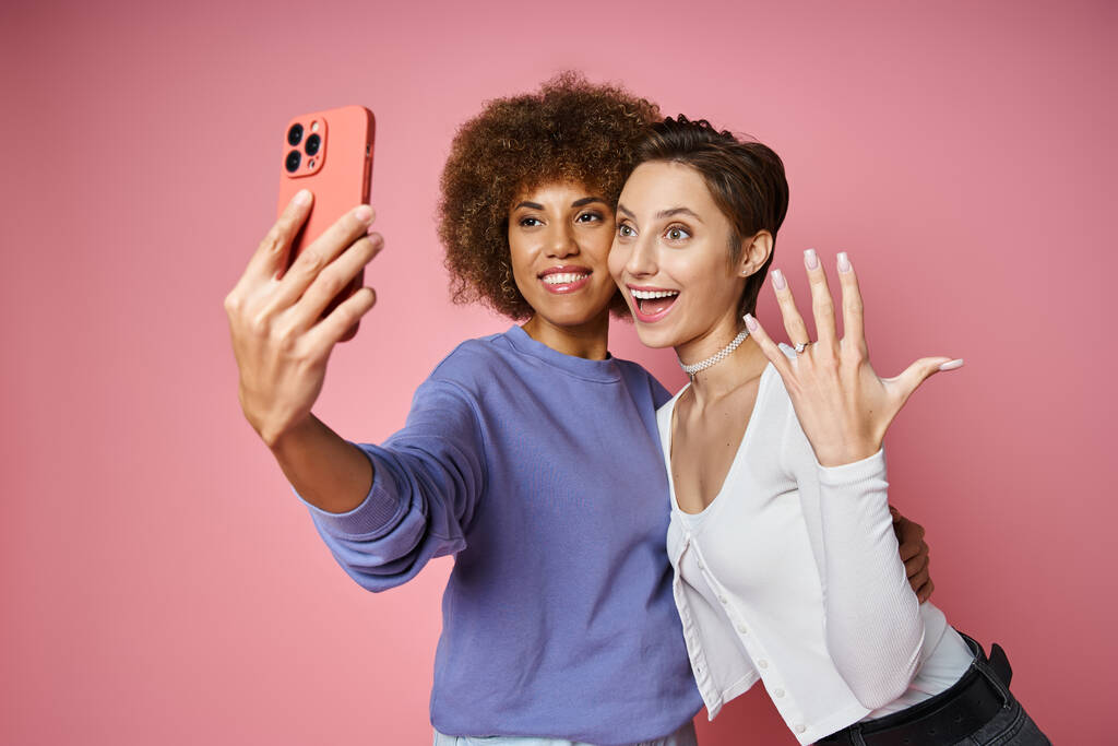 vrolijk lesbisch vrouw tonen verlovingsring op vinger terwijl het nemen van selfie met verloofde op roze - Foto, afbeelding
