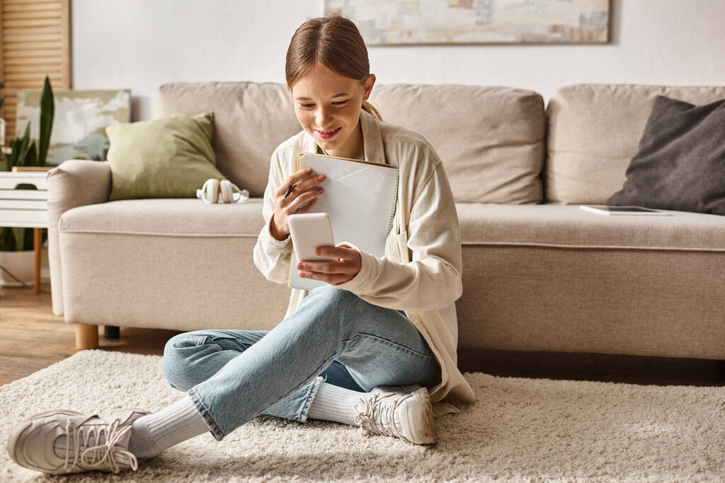 menina adolescente feliz segurando seu notebook e smartphone e sentado perto do sofá com fones de ouvido - Foto, Imagem