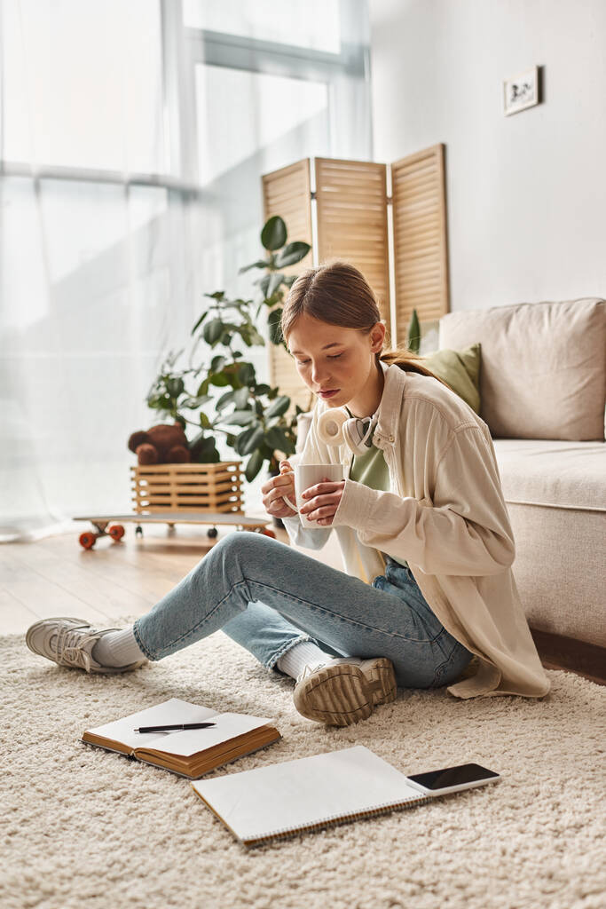Teen ragazza gode di una pausa caffè a casa e guardando smartphone e notebook sul tappeto - Foto, immagini