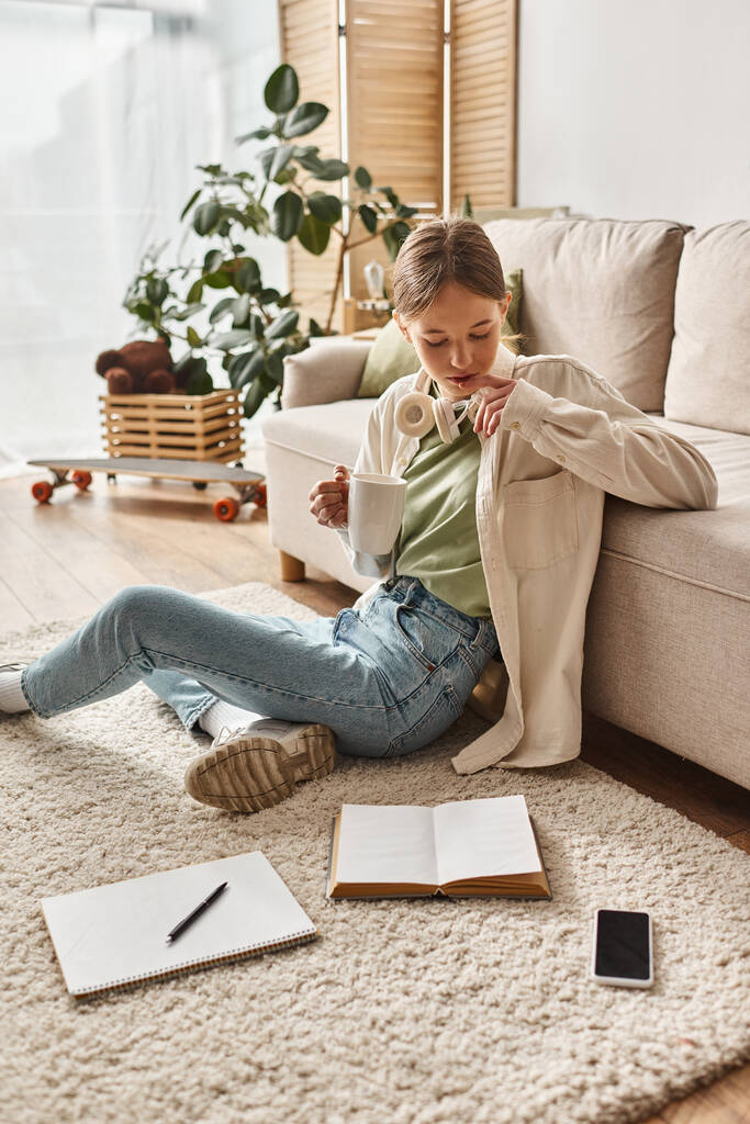 Девочка-подросток с наушниками наслаждается кофе дома и смотрит на смартфон и ноутбук на ковре - Фото, изображение