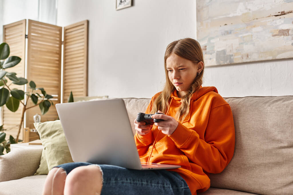 teen girl with joystick and laptop playing game and sitting on sofa at home, Σαββατοκύριακο δονήσεις - Φωτογραφία, εικόνα