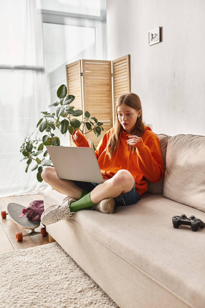 gen z adolescent fille souriant lors d'un appel vidéo sur un ordinateur portable et assis près joystick sur le canapé - Photo, image