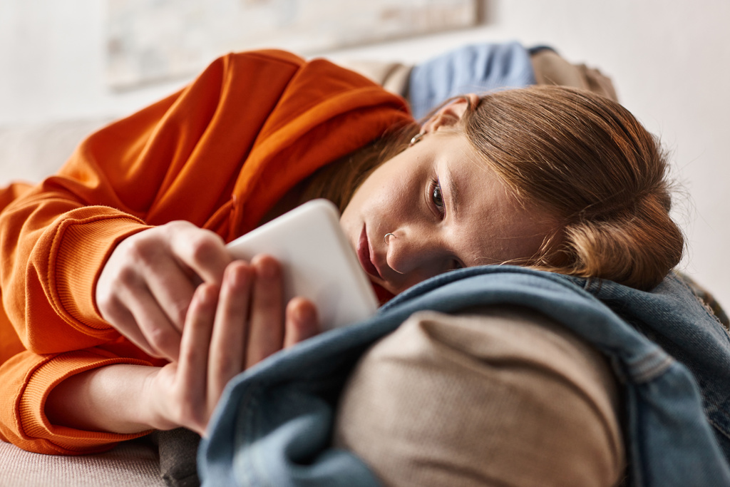 Unruhiges Teenager-Mädchen benutzt ihr Smartphone und fühlt sich im Wohnzimmer auf dem Sofa liegend - Foto, Bild
