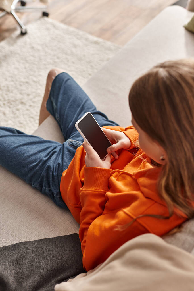 zoomer chica adolescente usando su teléfono inteligente y sentado en el sofá en la sala de estar, usuario de redes sociales - Foto, imagen