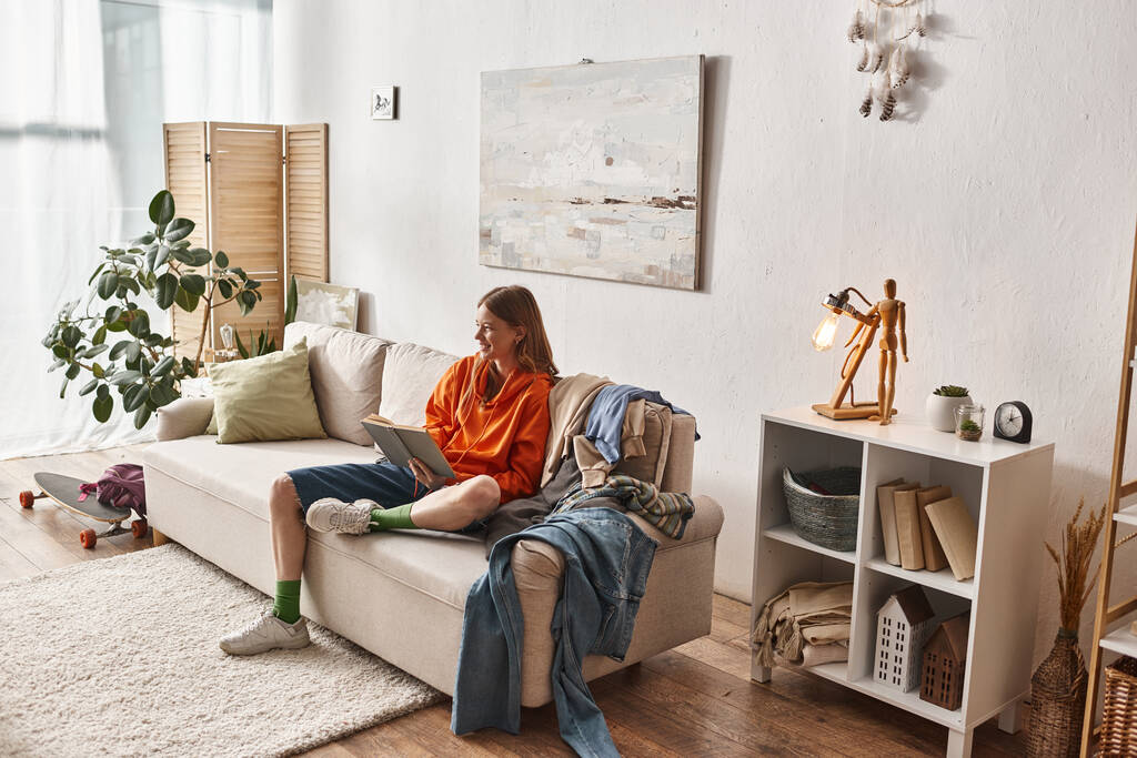 счастливая девочка-подросток читает книгу, сидя на диване рядом с кучей одежды в квартире - Фото, изображение