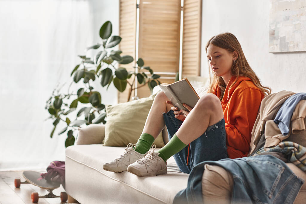 подросток занимается чтением книги и сидит на диване рядом с грязной кучей одежды в квартире - Фото, изображение