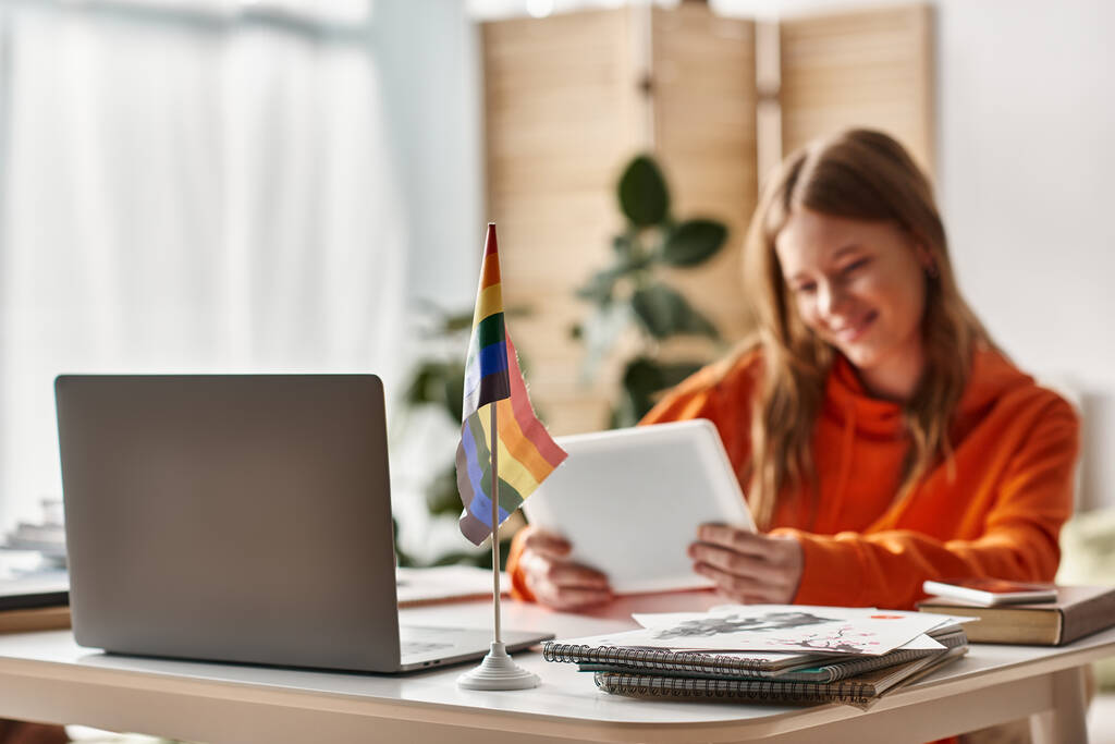 Веселая девочка-подросток с планшетом, участвующая в процессе электронного обучения возле гордого флага lgbtq на столе - Фото, изображение