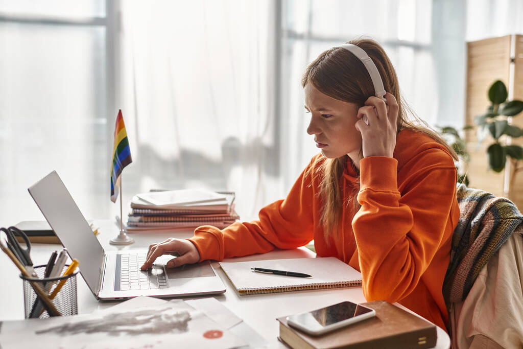 Концентрована дівчина-підліток у бездротових навушниках електронне навчання поруч з прапором гордості та канцелярськими товарами - Фото, зображення