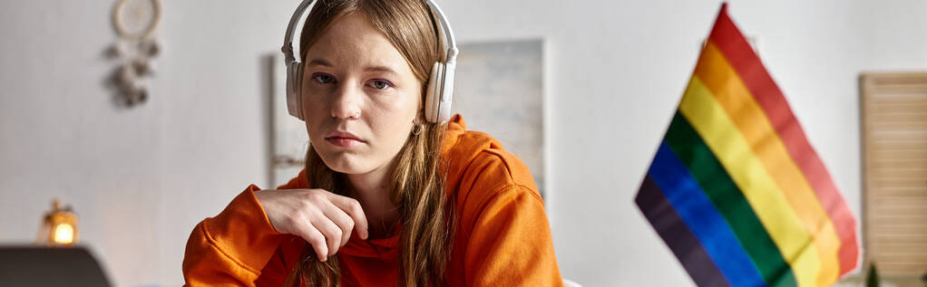 unavený mladý dospívající dívka v bezdrátových sluchátkách při pohledu na kameru vedle vlajky hrdosti, banner - Fotografie, Obrázek