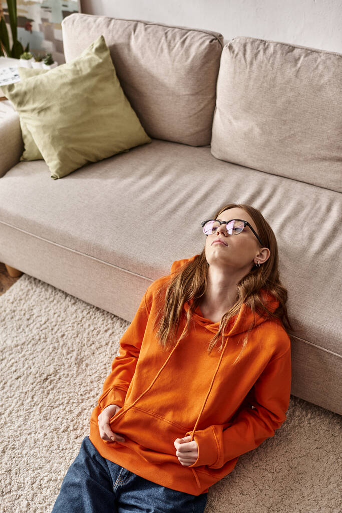 disgustada chica adolescente en naranja sudadera con capucha salones cerca de sofá en casa, melancolía y soledad de adolescente - Foto, Imagen