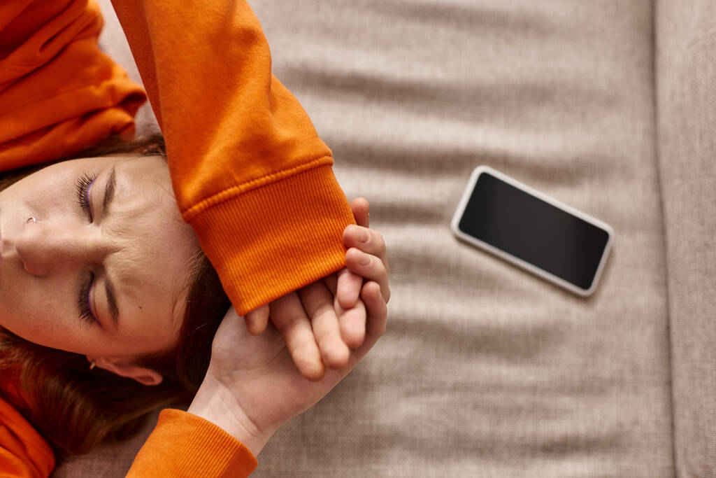 вид зверху дівчини-підлітка в помаранчевому светрі, що лежить біля смартфона на дивані, стрес від соціальних медіа - Фото, зображення