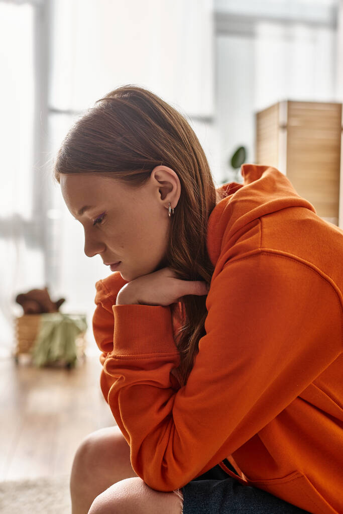 upset teenager girl in orange hoodie sitting on couch in Moden apartment, solitude and sadness - Photo, Image