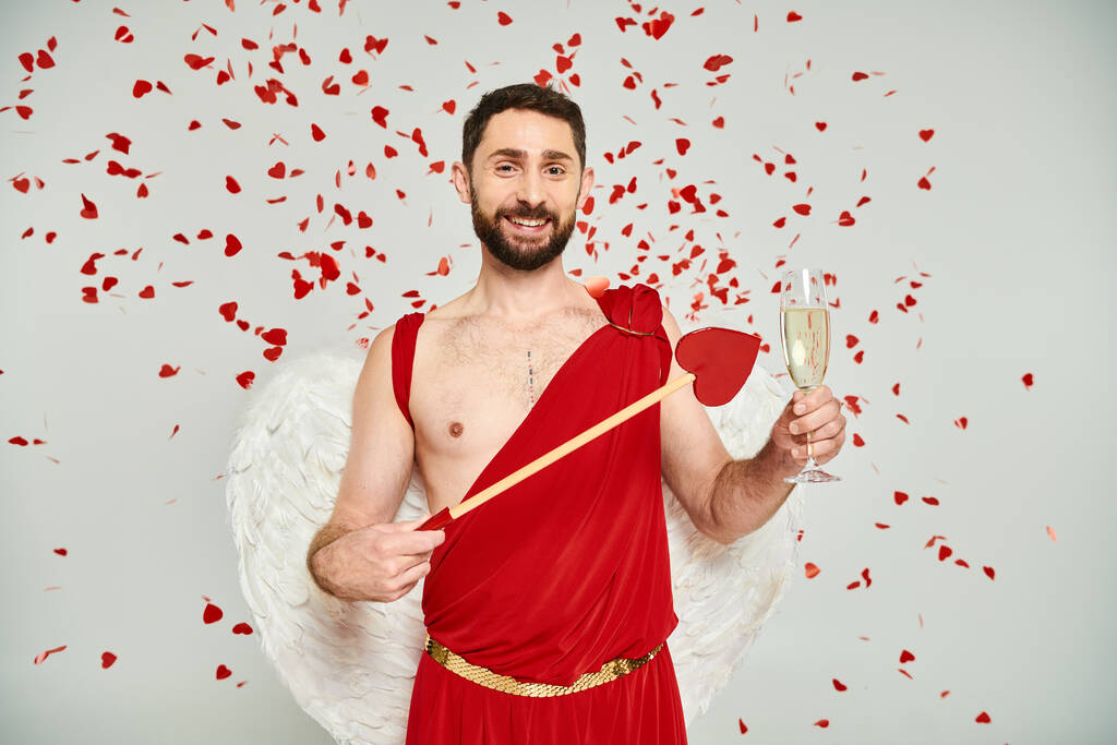 hombre barbudo en traje de Cupido con flecha en forma de corazón y champán bajo confeti rojo en gris - Foto, Imagen