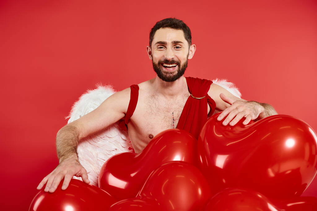 vousatý muž v amorfním kostýmu a křídlech, smějící se blízko balónků ve tvaru srdce na červeném, den svatého Valentýna - Fotografie, Obrázek