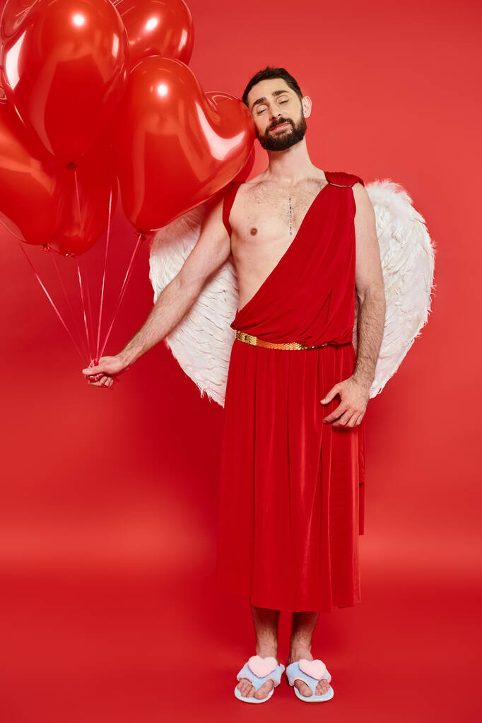 romántico barbudo hombre en traje de Cupido con los ojos cerrados y globos rojos en rojo, San Valentín día - Foto, Imagen