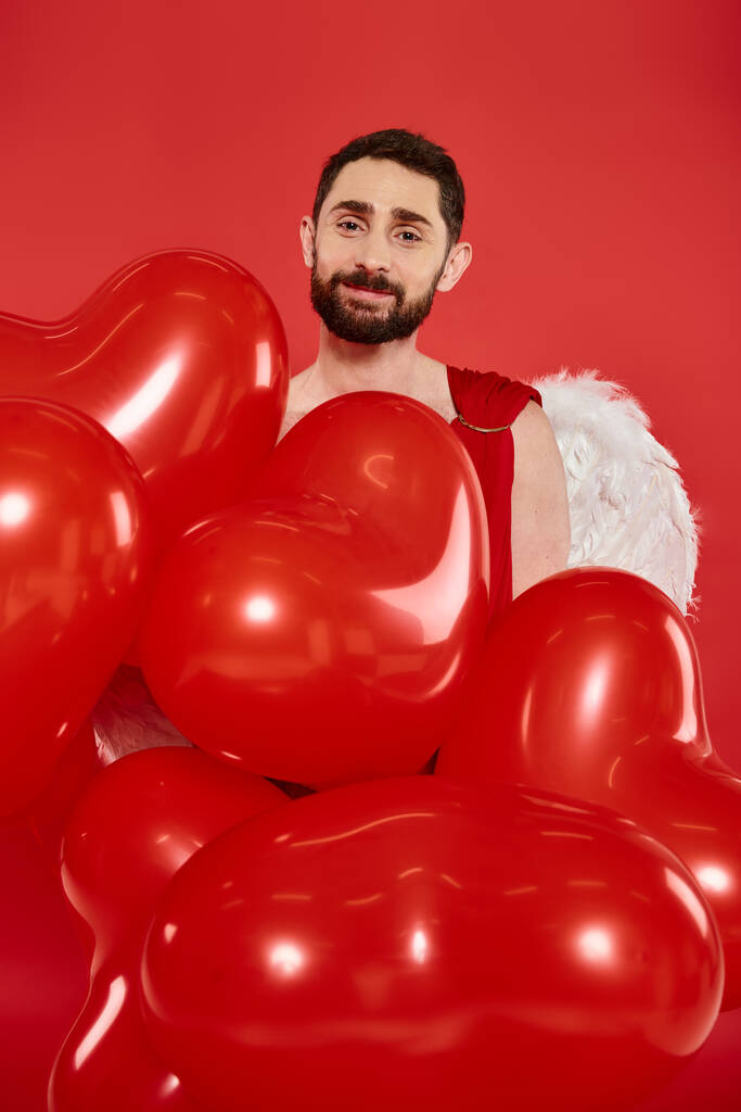 complacido barbudo hombre Cupido con globos en forma de corazón mirando a la cámara en rojo, San Valentín fiesta - Foto, imagen