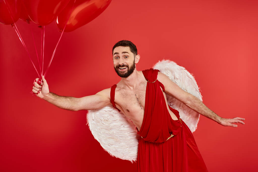 усміхнений бородатий чоловік в куполоподібному костюмі позує з повітряними кулями у формі серця на червоному, день святого Валентина - Фото, зображення
