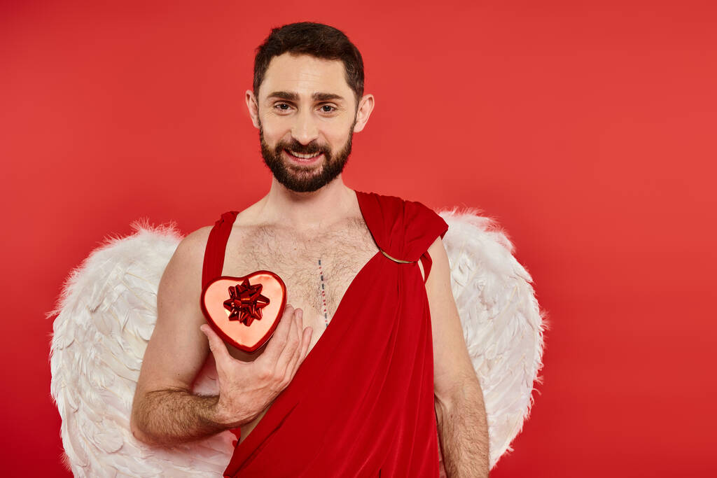 smiling bearded cupid man showing heart-shaped gift box on red, st valentines day themed event - Photo, Image