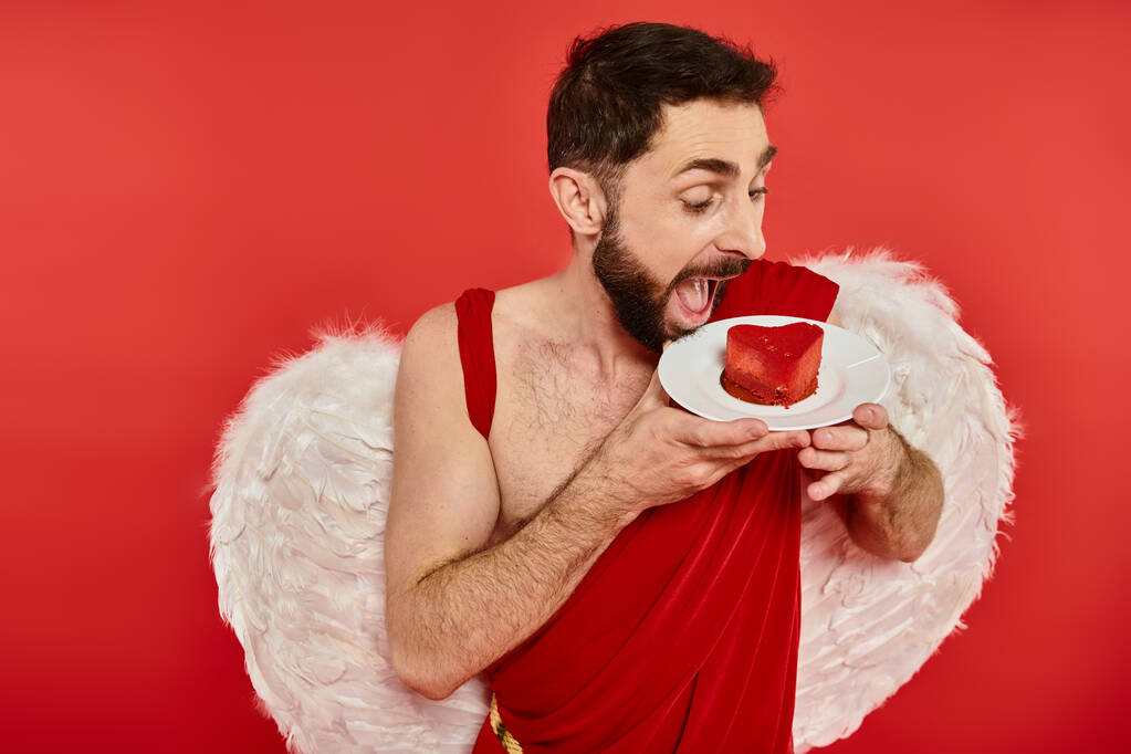 bearded cupid man with open mouth looking at delicious heart-shaped cale on red, st valentines treat - Photo, Image
