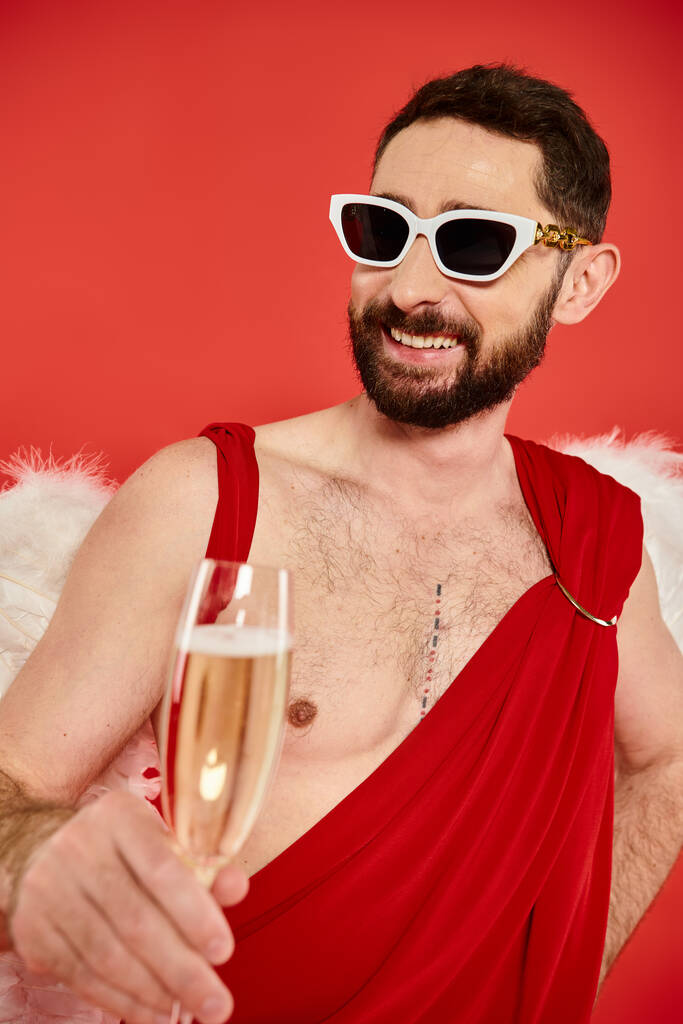 Güneş gözlüklü ve aşk tanrısı kostümlü sakallı adam kırmızı kadehle gülümsüyor, Sevgililer Günü. - Fotoğraf, Görsel