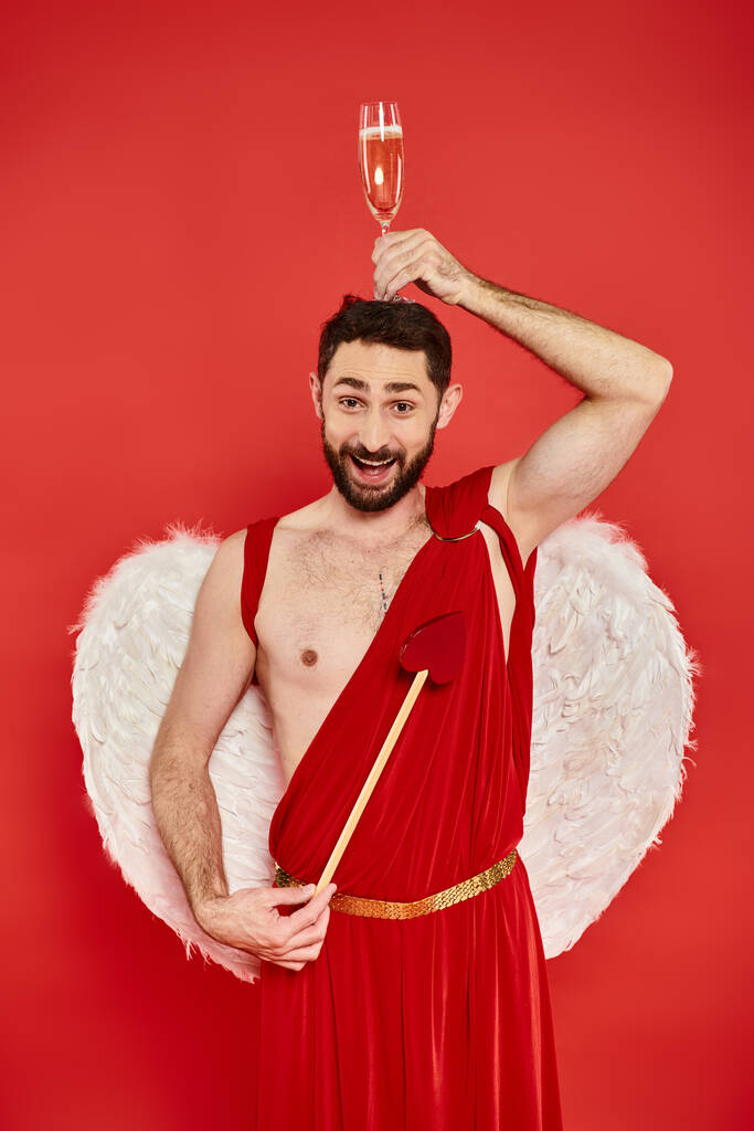 lustiger und fröhlicher Mann im Amor-Kostüm posiert mit herzförmigem Pfeil und Champagnerglas auf dem Kopf - Foto, Bild