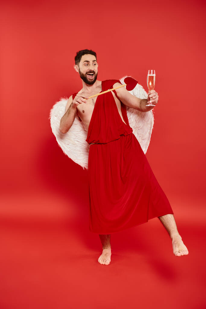 збуджений бородатий чоловік у костюмі з серцеподібною стрілкою та келихом шампанського на червоній, повній довжині - Фото, зображення