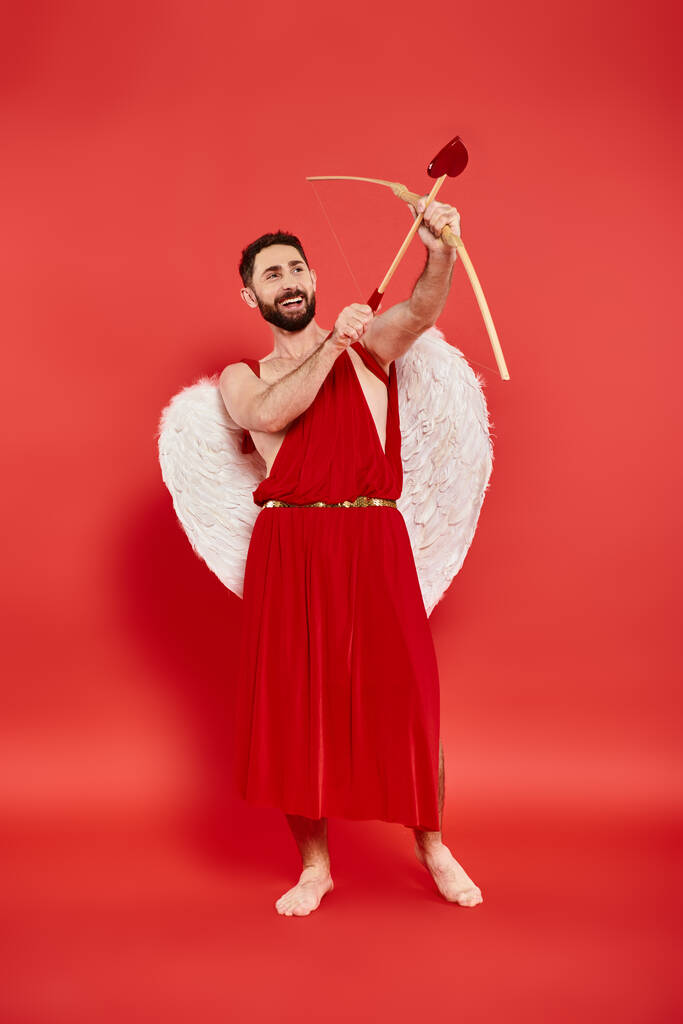 piena lunghezza di uomo eccitato in costume cupido tiro con l'arco sul rosso, concetto di San Valentino - Foto, immagini
