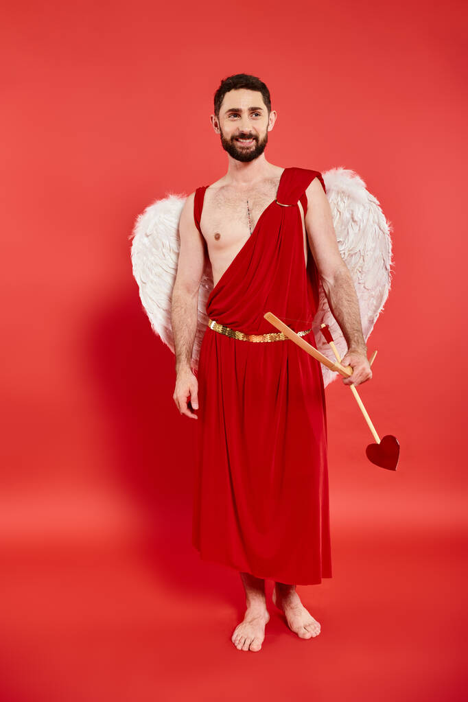 uomo barbuto sorridente a piedi nudi in costume cupido con arco e freccia in piedi e guardando lontano sul rosso - Foto, immagini