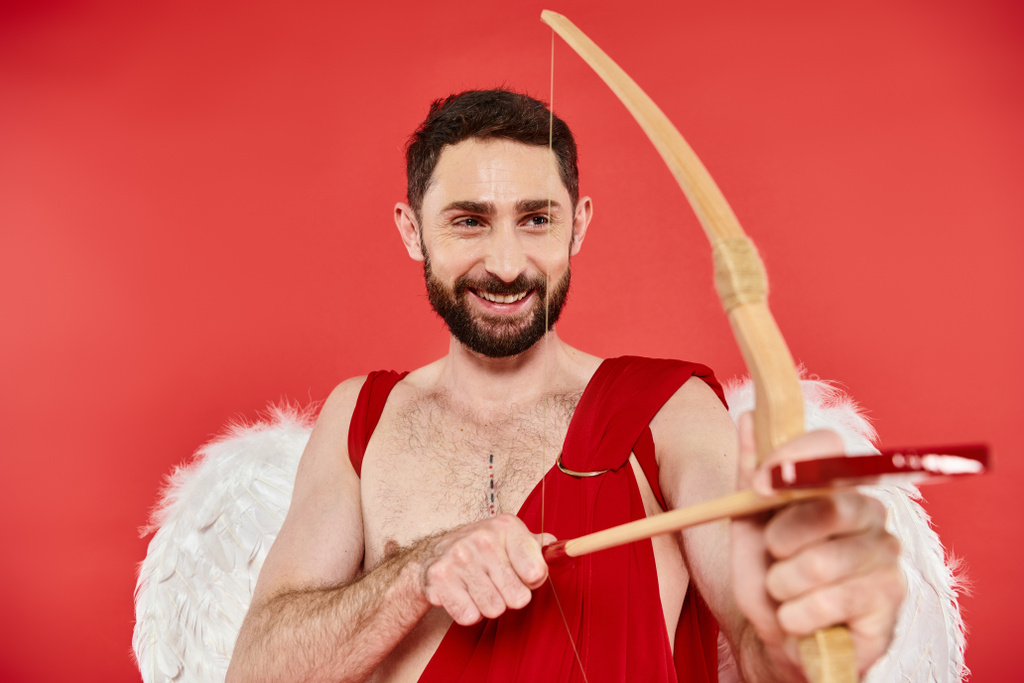 fröhlicher bärtiger Mann im Amor-Kostüm, der beim Zielen und Bogenschießen auf rote, - Foto, Bild
