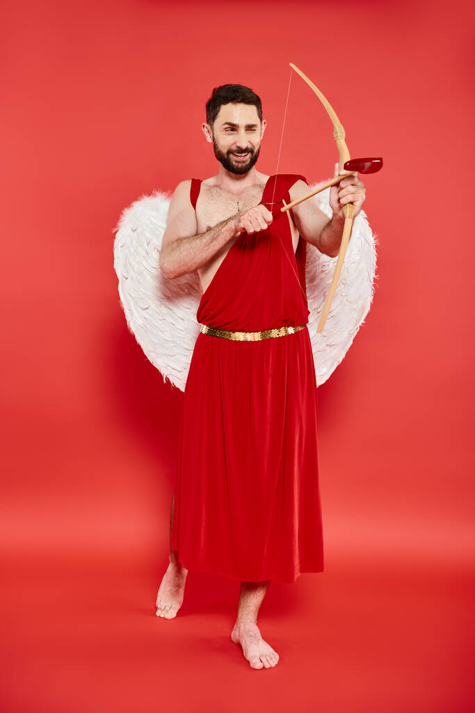 pleine longueur de sourire pieds nus homme en costume de Cupidon regardant loin et tir à l'arc sur fond rouge - Photo, image