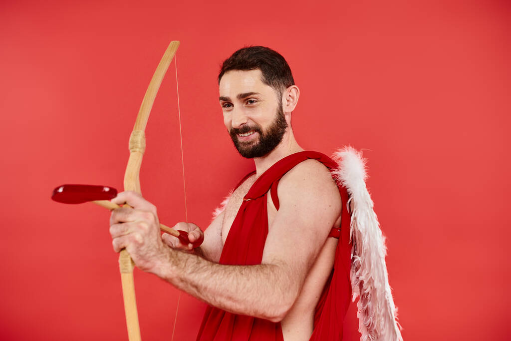 hombre barbudo feliz vestido como cupido arqueo con flecha en forma de corazón en rojo, día de San Valentín - Foto, Imagen