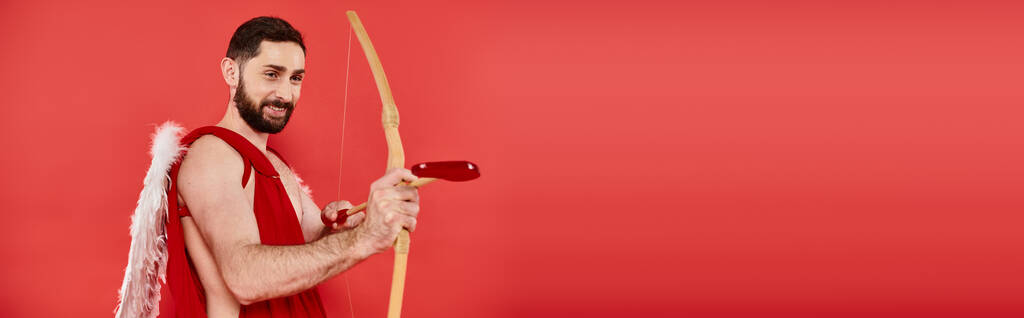 радісний бородатий чоловік, одягнений як купідон, що стріляє з лука зі стрілою у формі серця на червоному, банер - Фото, зображення
