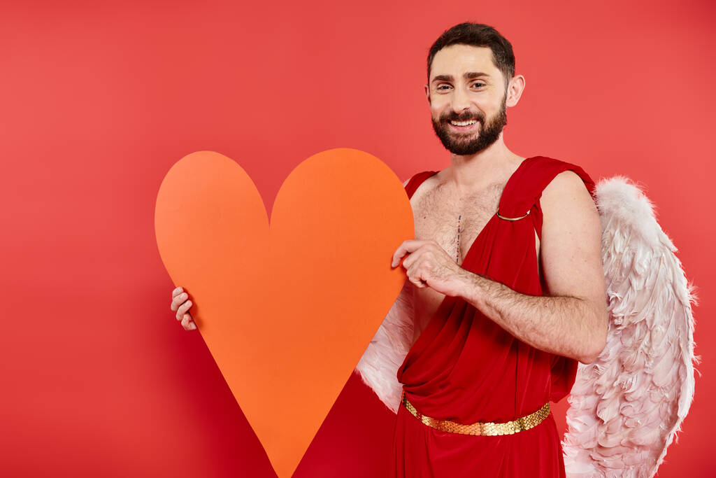 zadowolony brodaty mężczyzna w kostium Kupidyna gospodarstwa ogromny pomarańczowy papier serce na czerwono, święte Walentynki - Zdjęcie, obraz
