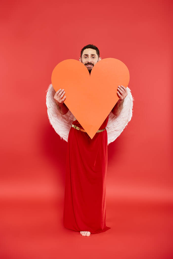 Lächelnder bärtiger Mann im Amor-Kostüm mit riesigem orangefarbenem Herz auf rotem Valentinstag-Kostüm - Foto, Bild