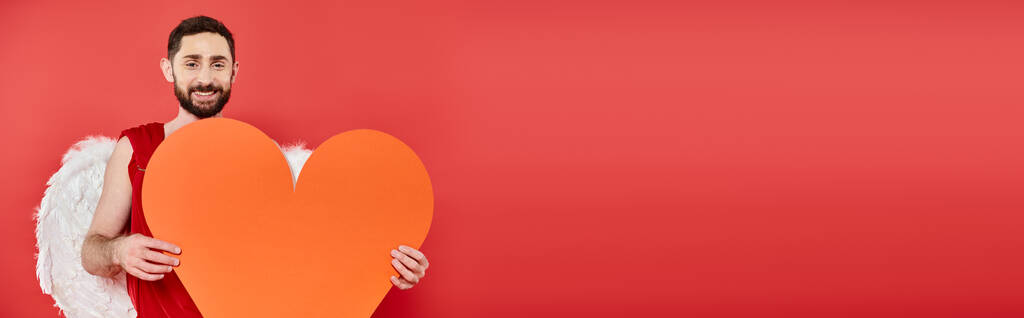 romantischer bärtiger Mann im Amor-Kostüm mit riesigem Papierherz auf rotem Grund, Valentinstag, Banner - Foto, Bild