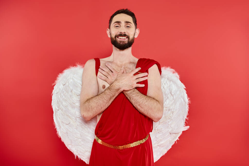 gelukkig bebaarde man in cupido kostuum staan met handen op de borst en kijken naar de camera op rood - Foto, afbeelding