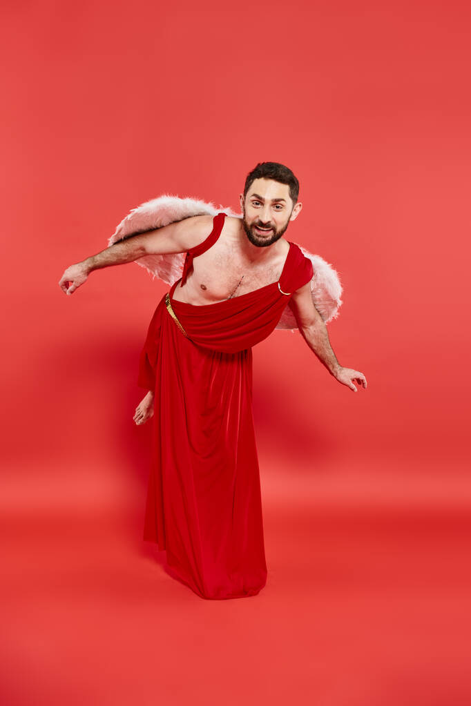 funny bearded man dressed as cupid bowing and looking at camera on red, st valentines themed event - Photo, Image