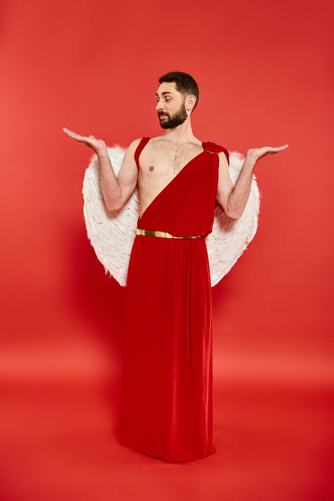 longitud completa del hombre vestido como Cupido mirando sus palmas abiertas en rojo, día de San Valentín - Foto, Imagen