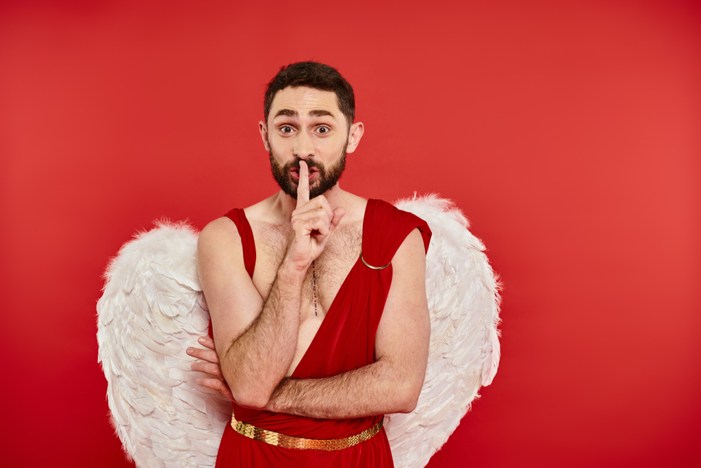Aşk tanrısı kostümü giymiş heyecanlı sakallı bir adam "şşş" işareti gösteriyor ve kırmızı sevgililer gününde kameraya bakıyor. - Fotoğraf, Görsel
