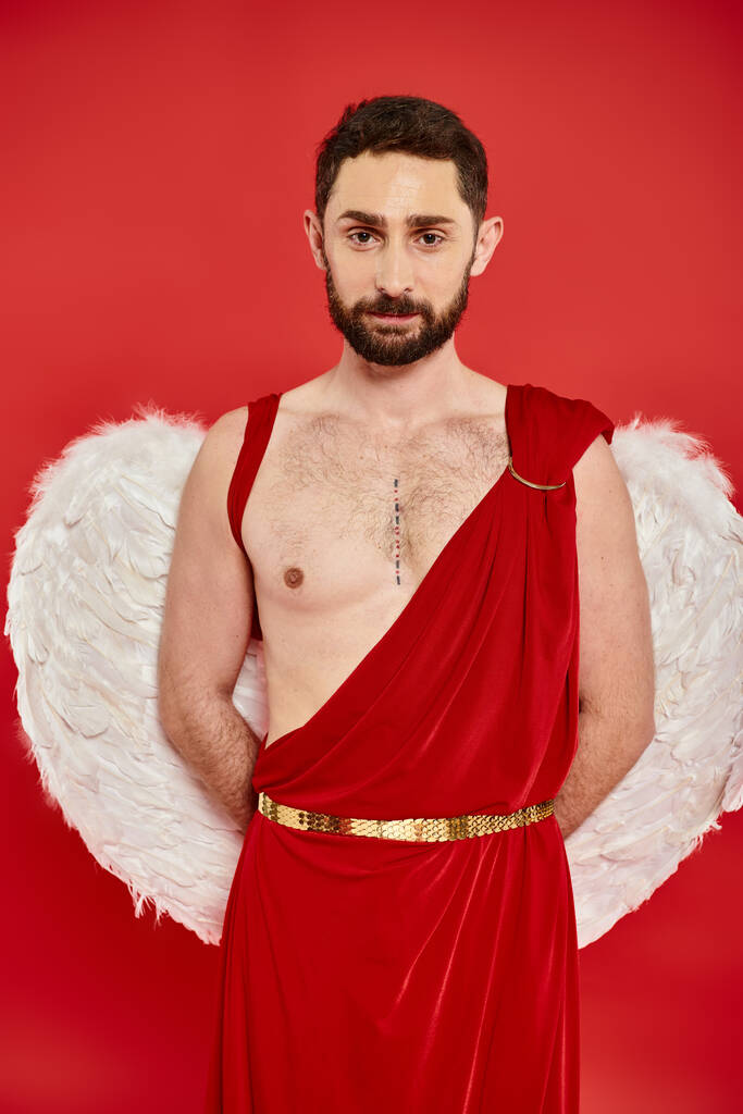 homme barbu en costume de Cupidon et ailes posant avec les mains derrière le dos et regardant la caméra sur rouge - Photo, image