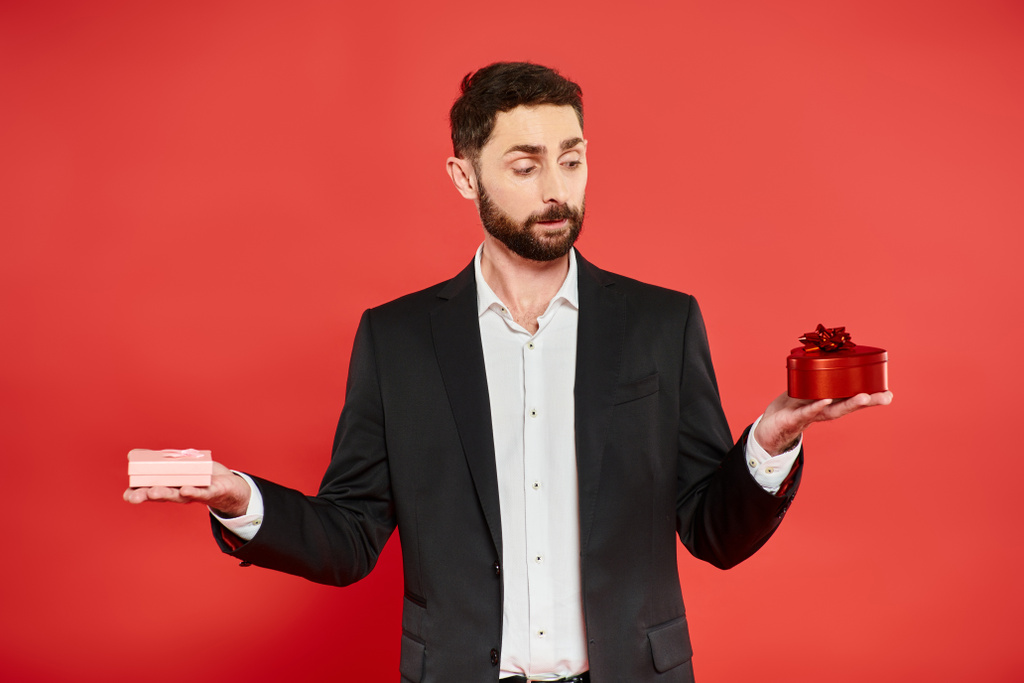 elegant bearded businessman in black suit choosing Saint Valentines day present on red backdrop - Photo, Image