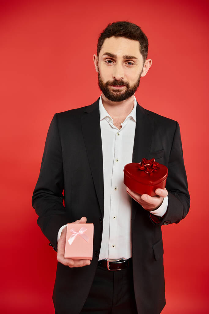 positiver bärtiger Mann im eleganten Anzug mit Valentinstagsgeschenken und Blick in die Kamera auf rot - Foto, Bild