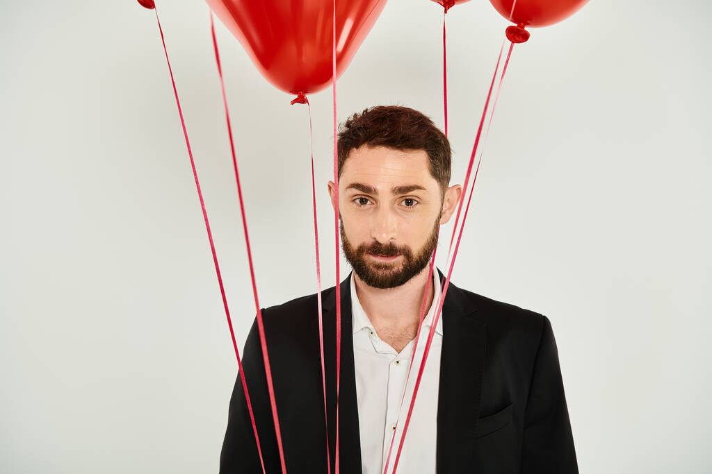 charismatique homme barbu regardant la caméra près de ballons festifs rouges sur gris, concept st valentines - Photo, image