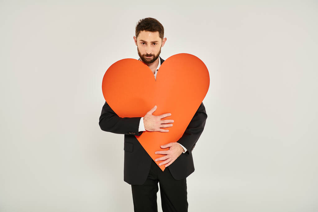 Sakallı, karizmatik bir adam kocaman kağıt kalbi kucaklıyor ve gri Sevgililer Günü 'nde kameraya bakıyor. - Fotoğraf, Görsel