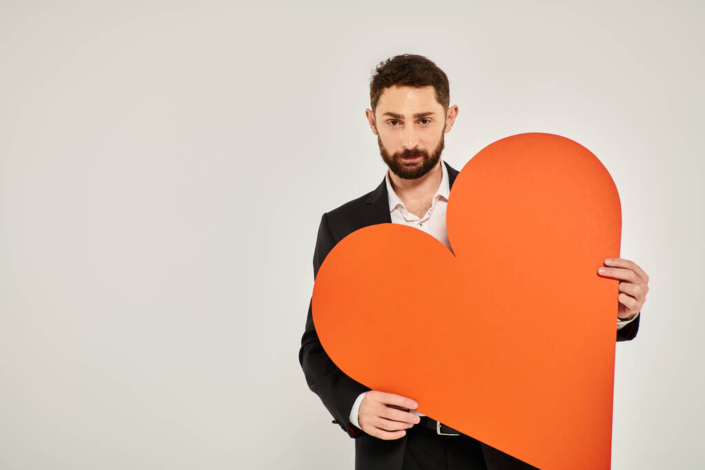 красивый элегантный мужчина с огромным бумажным сердцем, смотрящим в камеру на сером, концепция Святого Валентина - Фото, изображение